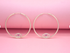 A pair of gold hoop earrings featuring sparkling evil eye charms with pink stone centers rests against a soft pink background.