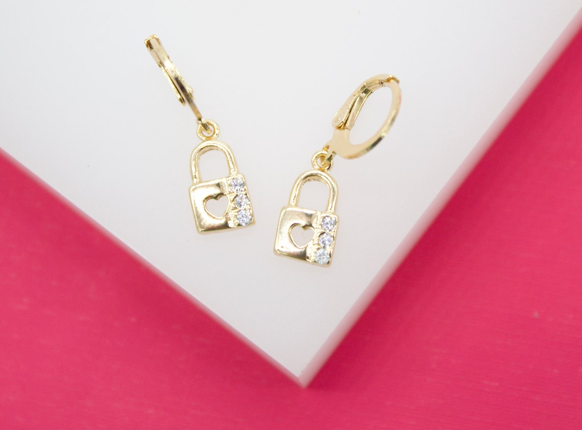 A pair of gold padlock drop earrings featuring a heart cutout and sparkling cubic zirconia stones rests on a white and pink display.