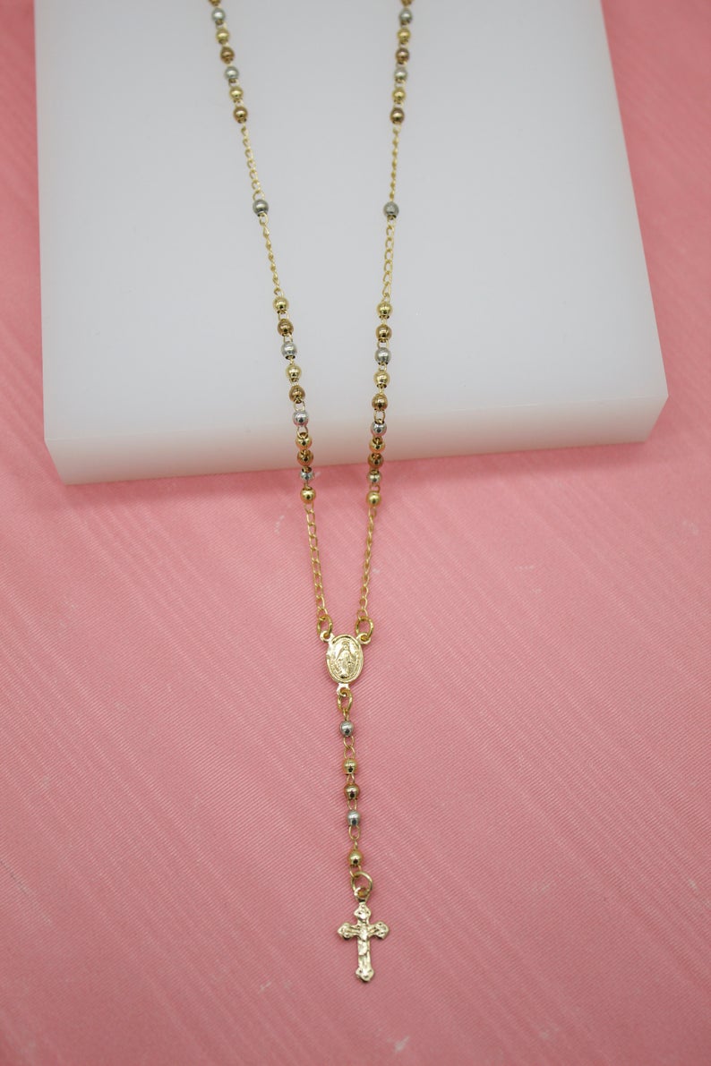 A delicate gold rosary necklace with alternating gold and silver-toned beads rests on a white block against a pink background.
