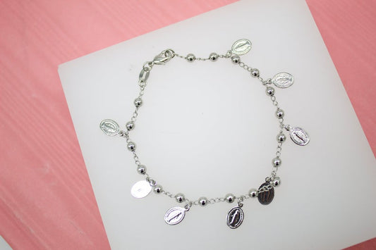 A delicate silver rosary bracelet featuring hanging miraculous medals rests on a white display against a pink background.