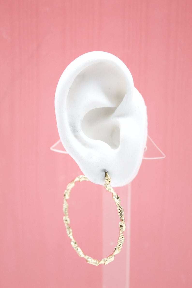A white mannequin ear displays a textured gold hoop earring against a pink background.