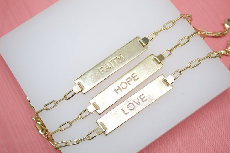 Three gold link chain bracelets with engraved bar pendants reading "Faith," "Hope," and "Love" rest neatly on a white display stand against a pink background.