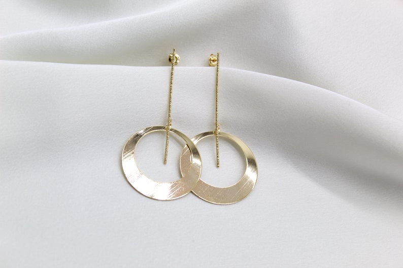 A pair of elegant gold drop earrings featuring slender textured posts and open circle pendants rest against a soft white draped fabric.