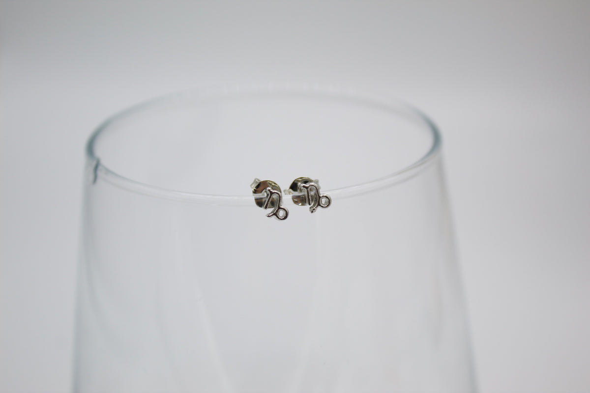 A pair of sterling silver Capricorn zodiac stud earrings rests on the rim of a clear glass.