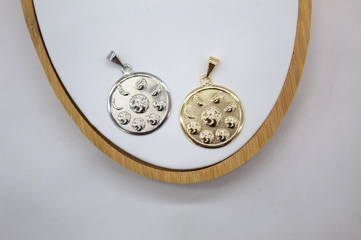 A silver and a gold celestial medallion pendant rest side-by-side on a round white display tray with a wooden rim.