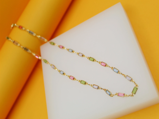 A delicate gold chain necklace featuring multi-colored rectangular crystal stones rests elegantly on a white display block against a vibrant yellow background.