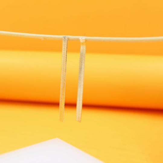 Two elegant flat gold snake chain earrings dangle from a thin twig against a vibrant yellow backdrop.