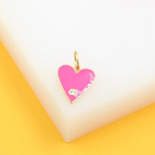 A gold heart-shaped pendant featuring vibrant hot pink enamel and a sparkling cubic zirconia stone rests on a white display block.