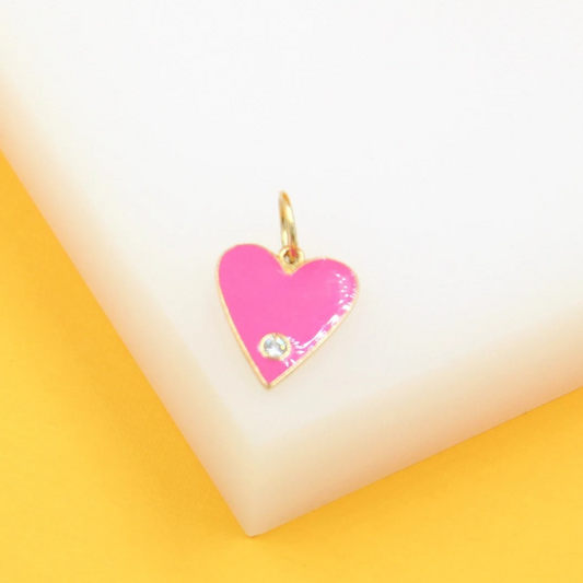 A gold heart-shaped pendant featuring vibrant hot pink enamel and a sparkling cubic zirconia stone rests on a white display block.