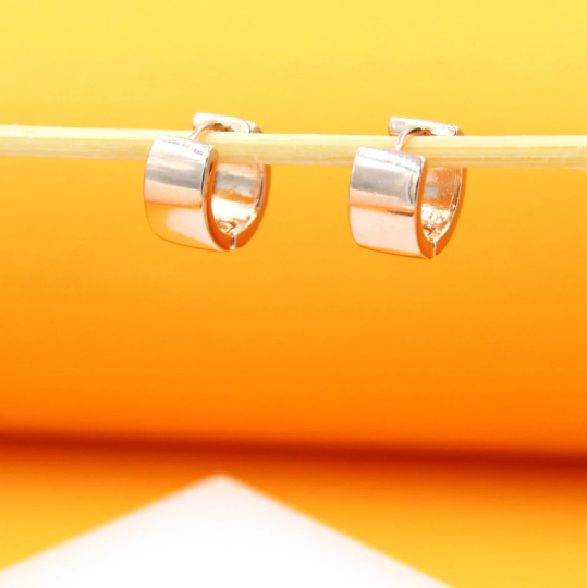 A pair of polished silver huggie hoop earrings hangs neatly from a thin wooden stick against a bright yellow and orange gradient backdrop.