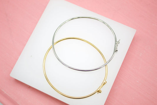 A silver and a gold bangle bracelet rest on a white display block against a pink background.