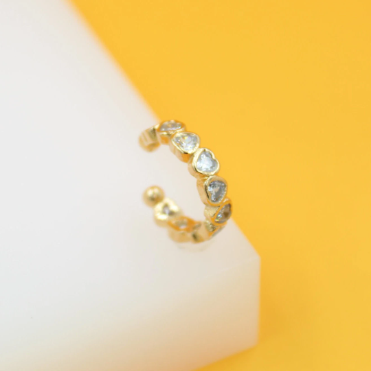 A gold ear cuff featuring sparkling heart-shaped crystals rests on a white display surface against a vibrant yellow backdrop.