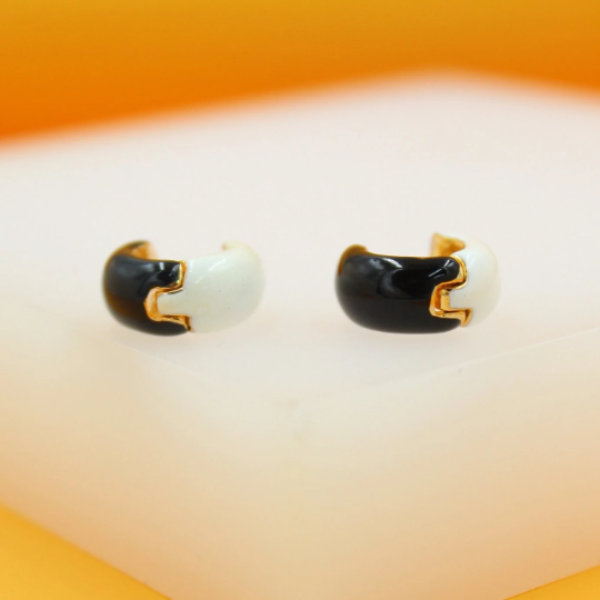 A pair of gold-plated huggie hoop earrings featuring a chic split design of glossy black and white enamel rest on a white display surface.
