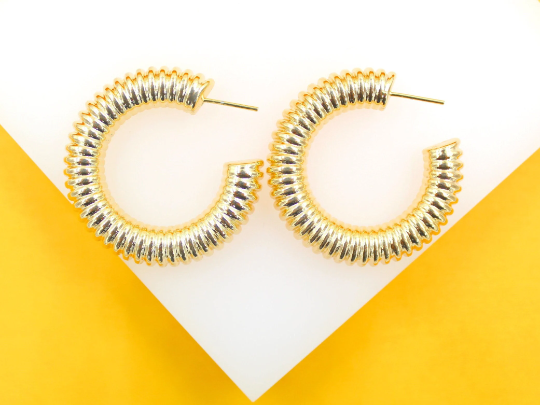A pair of gold-plated chunky ribbed hoop earrings rests against a white and yellow background.