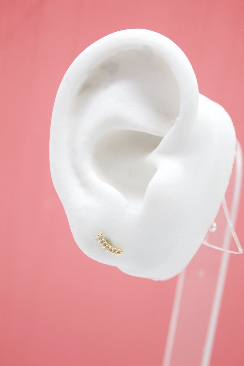 A curved gold cartilage earring with sparkling crystal stones rests on a white mannequin ear against a soft pink background.