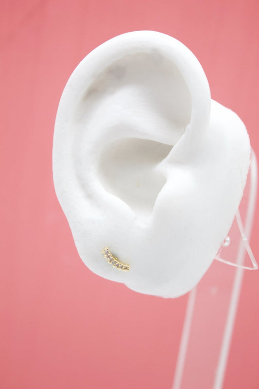 A curved gold cartilage earring with sparkling crystal stones rests on a white mannequin ear against a soft pink background.