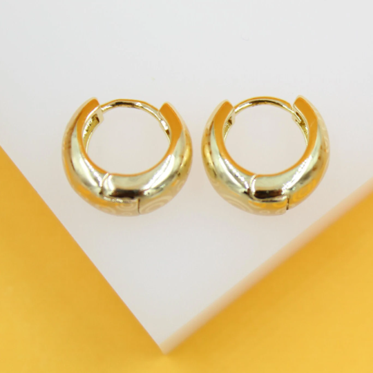 A pair of shiny gold huggie hoop earrings with subtle swirling patterns rests on a white display stand.
