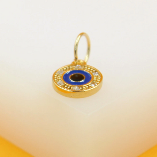 A small gold evil eye charm pendant rests on a white surface, featuring a blue enamel center and sparkling cubic zirconia accents.