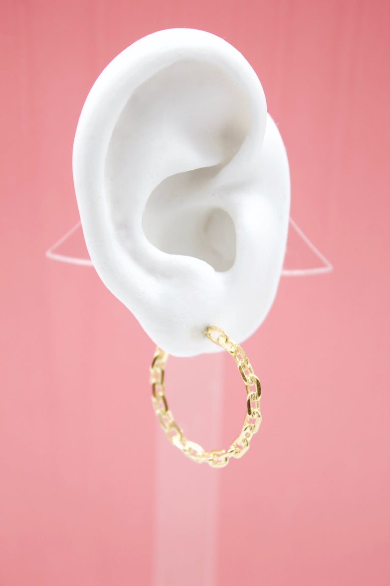 A white mannequin ear displays a textured gold chain link hoop earring against a pink background.