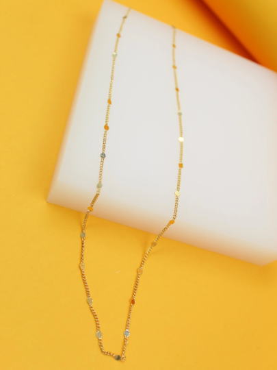 A delicate gold chain necklace with small circular coin charms drapes elegantly across a white display stand against a vibrant yellow background.
