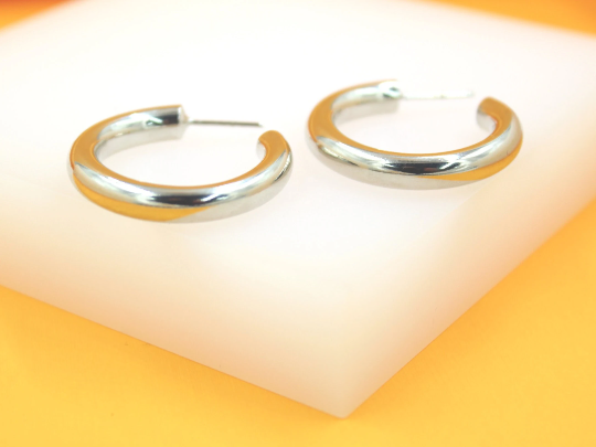 A pair of sleek silver hoop earrings rests neatly on a white display surface against a vibrant orange background.