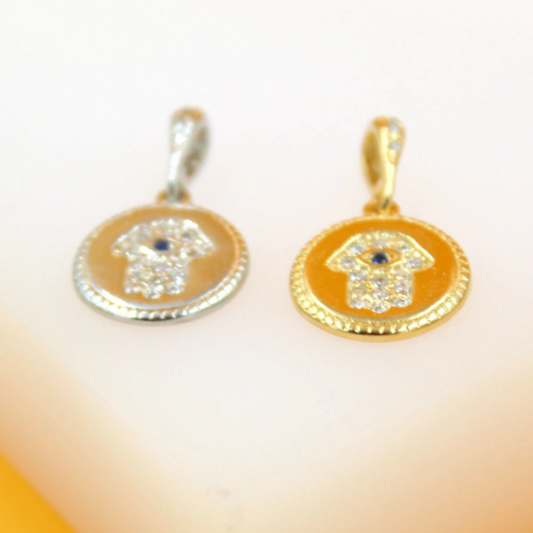 Two round Hamsa hand pendants with evil eye details rest on a surface, one in silver and the other in gold.
