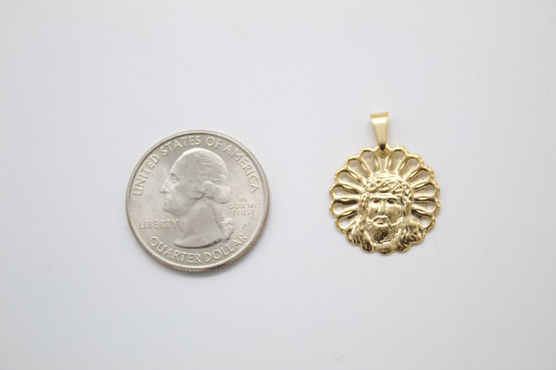 A gold Jesus face pendant rests next to a US quarter coin for size comparison on a light surface.