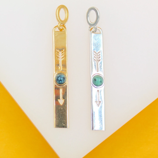 Two rectangular arrow-shaped pendants in gold and silver, featuring inlaid turquoise stones, stand upright against a plain background.