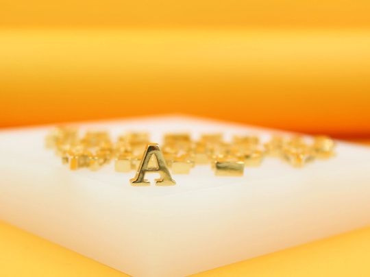 A shiny gold letter A charm stands prominently on a white display block with a bright yellow background.