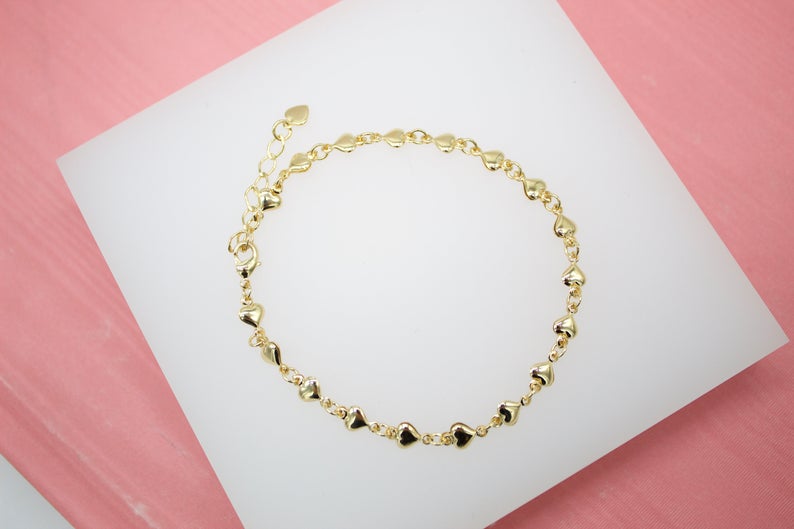 A delicate gold heart-link bracelet rests on a white display surface against a pink background.