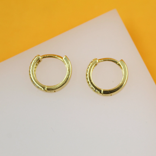 A pair of delicate gold huggie hoop earrings featuring tiny sparkling CZ stones rests on a white display surface against a vibrant orange background.