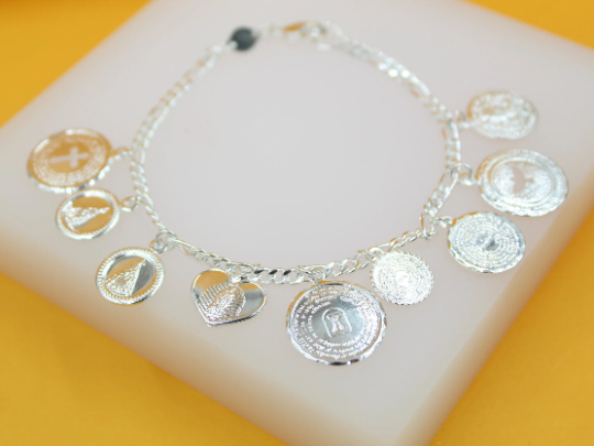 A delicate silver charm bracelet rests on a white display stand against a vibrant yellow background, showcasing multiple religious and symbolic charms.
