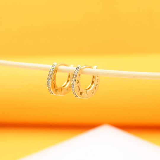 Two gold hoop earrings featuring celestial moon and star motifs hang delicately from a white stick against a warm yellow background.