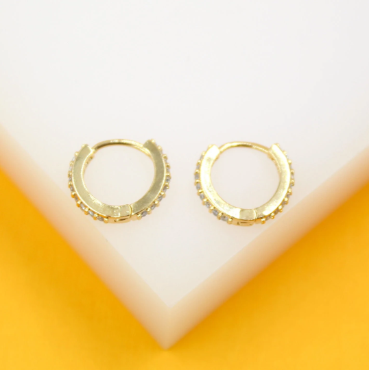 A pair of gold huggie hoop earrings embedded with small cubic zirconia stones rests elegantly on a white and yellow display stand.