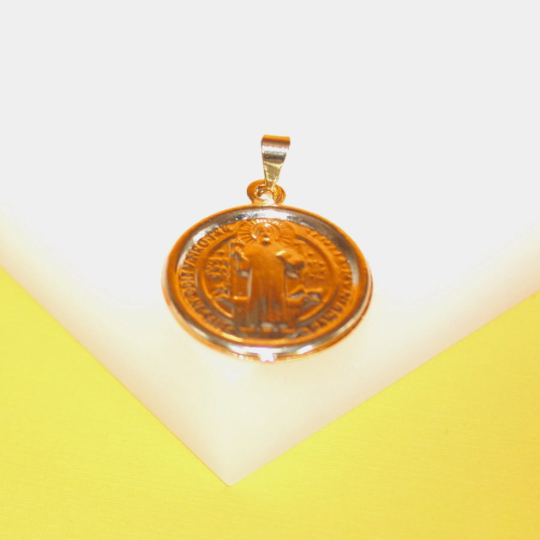 A round gold-plated Saint Benedict pendant rests on a bright yellow and white surface.