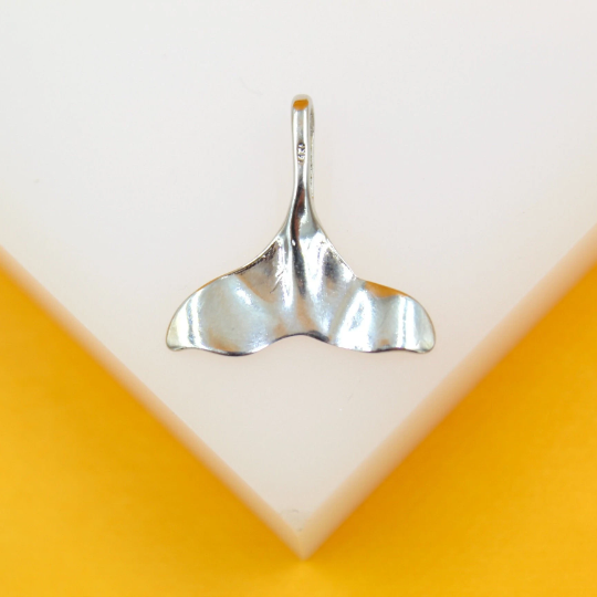 A silver whale tail pendant rests on a white display surface against a vibrant yellow background.