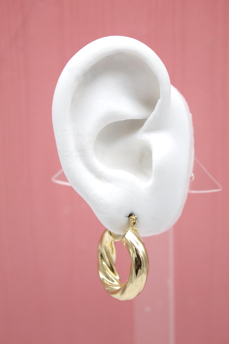 A white mannequin ear displays a shiny, thick twisted gold hoop earring against a soft pink background.