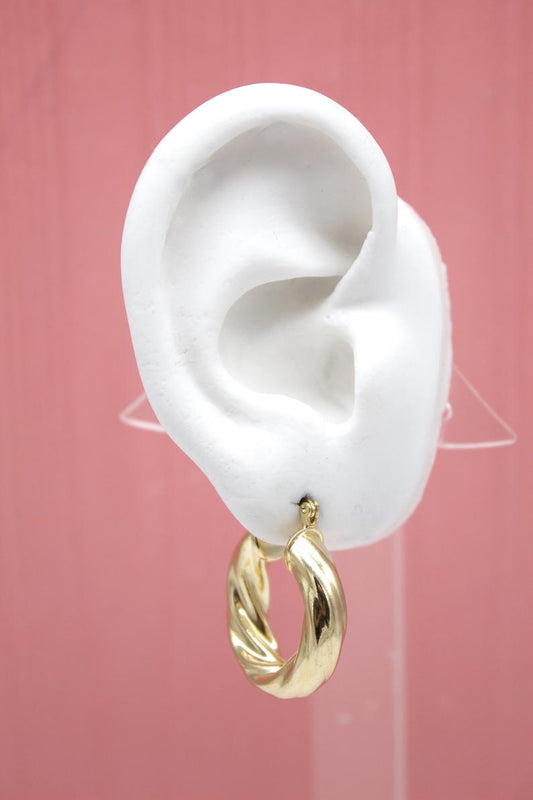 A white mannequin ear displays a shiny, thick twisted gold hoop earring against a soft pink background.