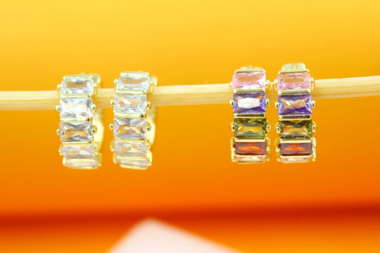 Two pairs of gold hoop earrings encrusted with baguette-cut multicolored and clear cubic zirconia stones hang from a wooden stick against an orange backdrop.
