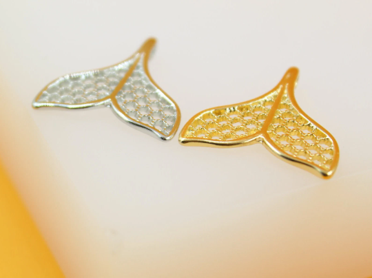 Two filigree mermaid tail charms in silver and gold rest on a light surface.
