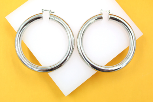 A pair of large, thick silver hoop earrings rests on a white square display against a vibrant yellow background.
