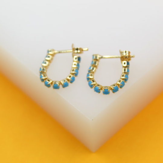 A pair of gold huggie hoop earrings embedded with small turquoise stones rests on a white display block against an orange background.