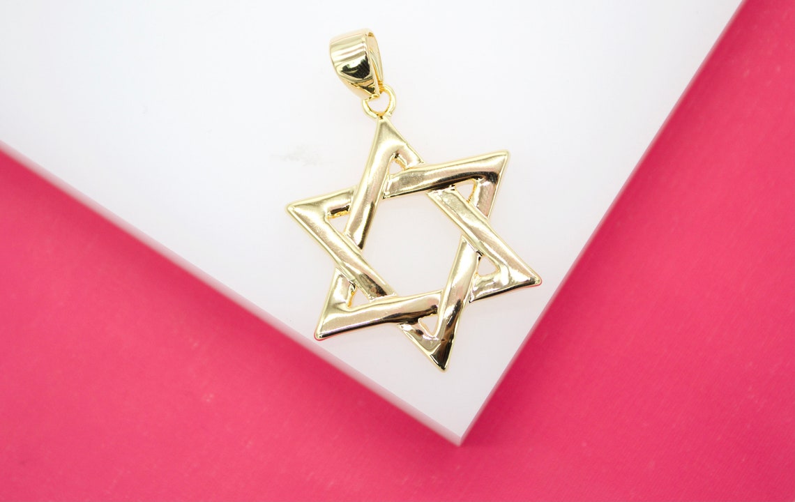 A polished gold Star of David pendant rests on a white display surface against a vibrant pink background.