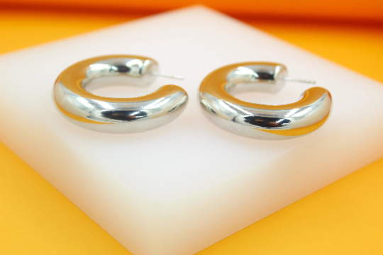 A pair of thick silver hoop earrings rests on a white display block against an orange background.
