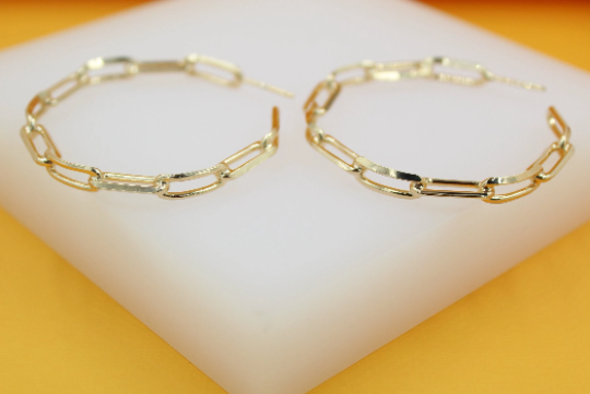 A pair of stylish gold paperclip chain hoop earrings rests on a white display surface.