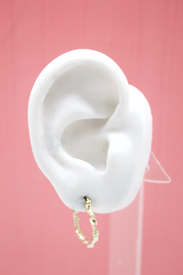 A white mannequin ear displays a stylish textured gold hoop earring against a pink background.