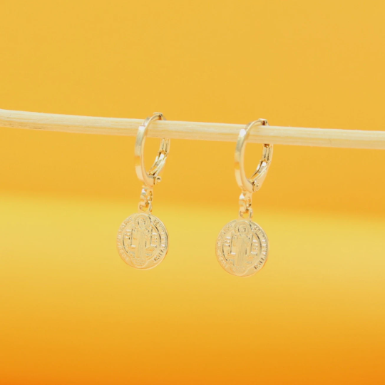 Two gold Saint Benedict medallion huggie hoop earrings hang delicately from a wooden stick against a bright yellow background.