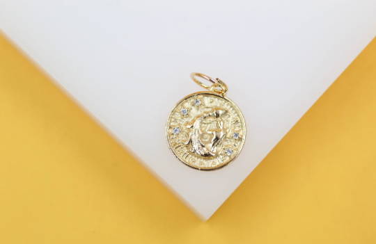 A round gold medallion pendant featuring an engraved astrological sign and sparkling cubic zirconia stones rests against a white and yellow background.