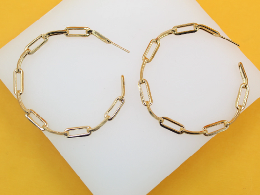 A pair of shiny gold paperclip chain hoop earrings rests on a white display against a vibrant yellow background.