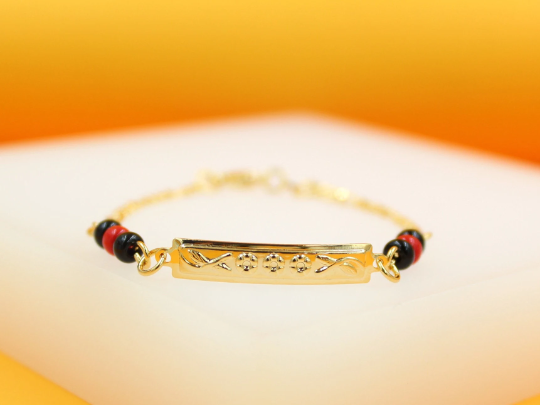 A gold ID bracelet rests on a white surface, featuring alternating black and red beads framing an engraved rectangular metal plate.
